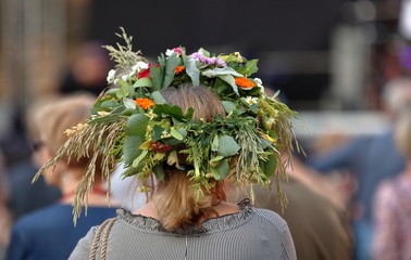 Kobieta, blond włosy, stoi tyłem (widoczna głowa i część ramion), w roślinnym, kwietnym, kolorowym wianku na głowie, w tle rozmyty tłum ludzi
