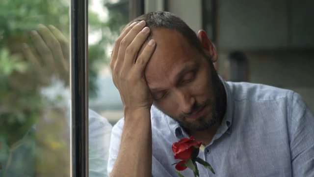 Sad, unhappy man holding red rose sitting by window
