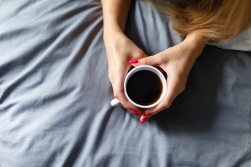 Top view of a Young girl in a white shirt holding a mug of coffee lying on the bed . Young woman drinking coffee at home in her bed, start, concept in the morning, early in the morning.