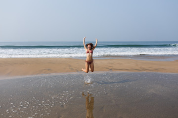 Slender young girl in a swimsuit is jumping on the sandy shore of the ocean. Happy Days. The concept of freedom, recreation, vacation. Flight of joy and fun. Reflection in water.