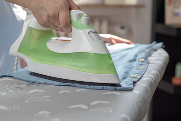 Woman is ironing a blue shirt with a green iron on the ironing board