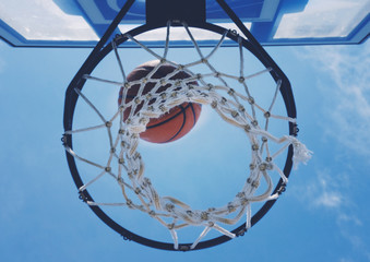 Basketball draining through hoop basket, shown through net looking up at sky.  Sport background for team or game.