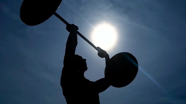 strong man is lifting a barbell over his head standing outdoor in sunny day,his silhouette against the sun