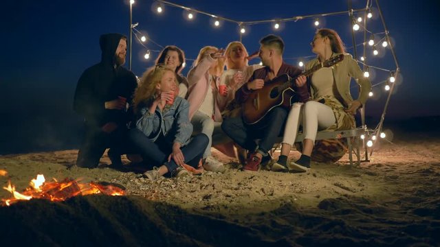 youth beach entertainment, happy people playing guitar and singing sitting by bonfire in sand on embankment in illumination of garlands