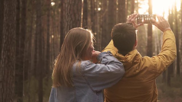 Young couple making selfie at smartphone at background of sunny green forest. Hiking emotionally concept of travelers spending weekend in wild side. Enjoyment, friendly moments, funny time outdoor