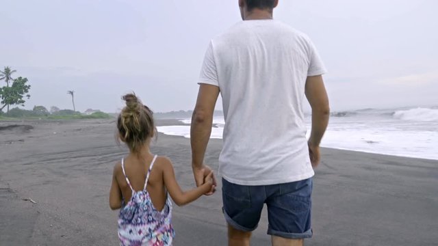 Rear view of man holding crab and walking with little daughter along ocean beach while having family vacation