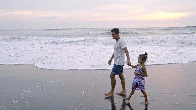 Side view of lovely little girl in bright summer dress holding hands with dad and walking along picturesque ocean beach at sunrise