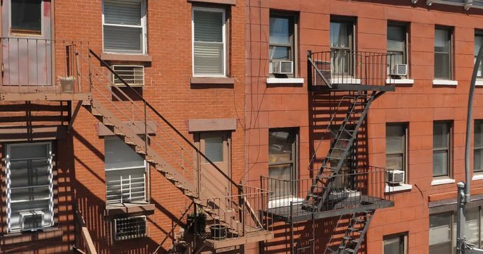 A daytime exterior establishing shot of a typical red brick apartment building with fire escapes along the side.  	