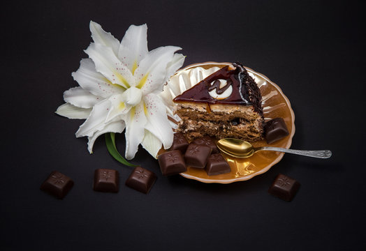 cut piece of chocolate cake on a yellow pearl plate with pieces of chocolate tiles on a black background and a lily flower