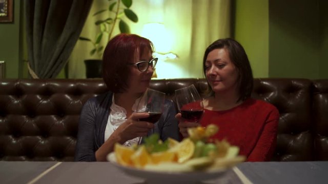 Two women sitting at a table in the restaurant talking clink glasses and drink red wine.