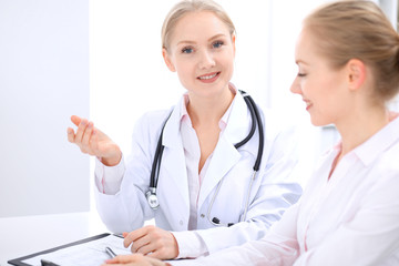 Fototapeta premium Blonde female doctor and patient talking in hospital office. Health care and client service in medicine