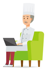 An elderly male chef wearing a cook coat is sitting on a sofa and operating a laptop computer