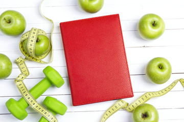 Red notepad with green dumbbells and apples with measuring tape on white wooden background. Sport,...