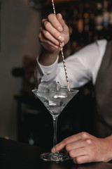 A vertical image of barman's hands stirring up ice cubes in a martini glass.