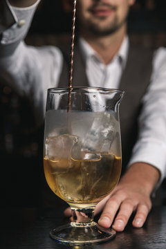 A vertical image of barman's hands stirring up a cocktail with ice cubes in a mixing glass.