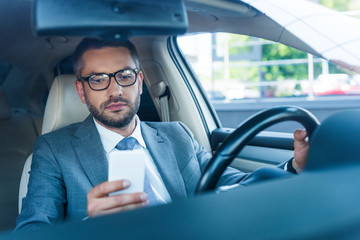 portrait of businessman in eyeglasses using smartphone in car