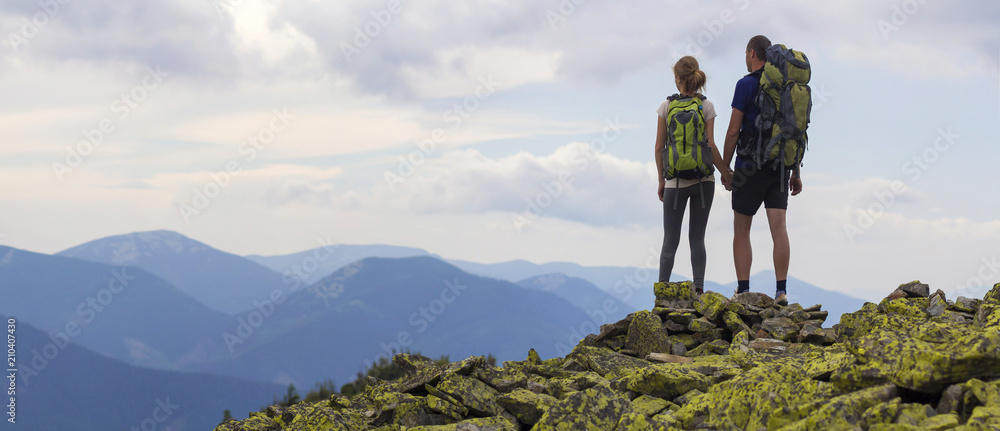 Wall mural Back view of young tourist couple with backpacks, athletic man and slim girl stand holding hands on rocky mountain top enjoying mountain panorama. Tourism, traveling and healthy lifestyle concept. - Wall murals