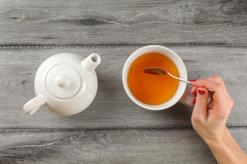 Table top view - woman hand holding spoon, stirring tea in white porcelain cup. - obrazy, fototapety, plakaty