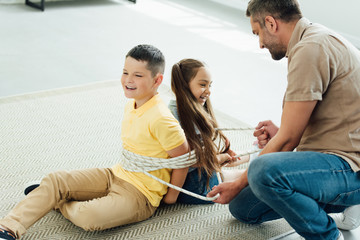 father tying smiling children with rope at home