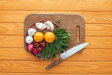 top view of fresh raw vegetables in plate on wooden cutting board and knife on wooden table top