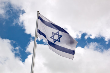Fototapeta premium Flag of Israel waving in the wind on flagpole against the sky with clouds on sunny day, close-up