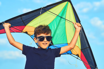 Happy boy in sunny glasses launches a bright kite at the seashore against a blue sky close up at sunset