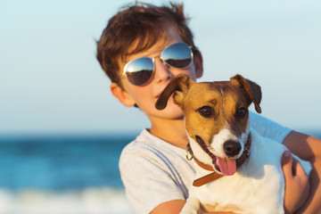 Happy boy hugging his dog breed Jack russell at the seashore against a blue sky close up at sunset. Best friends rest and have fun on vacation, play in the sand against the sea - obrazy, fototapety, plakaty