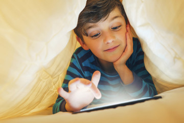 Happy smiling boy sits in bed under a blanket and plays on a smartphone in a game in the dark. The child's face is illuminated by a bright monitor - obrazy, fototapety, plakaty