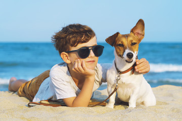 Happy 9 year old boy hugging his dog breed Jack russell at the seashore against a blue sky close up at sunset. Best friends rest and have fun on vacation, play in the sand against the sea - obrazy, fototapety, plakaty