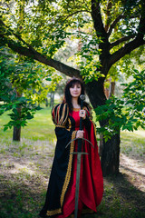 Obraz na płótnie Canvas The Queen with a sword in her hands. Beautiful girl in the crown. Fantasy. Medieval black and red dress with gold embroidery.