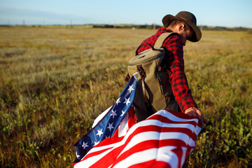 4th of July. American Flag.Patriotic holiday. Traveler with the flag of America. The man is wearing a hat, a backpack, a shirt and jeans. Beautiful sunset light. American style. 