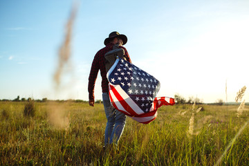 4th of July. American Flag.Patriotic holiday. Traveler with the flag of America. The man is wearing a hat, a backpack, a shirt and jeans. Beautiful sunset light. American style. 