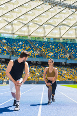 young athletic couple warming up legs before jogging on running track at sports stadium