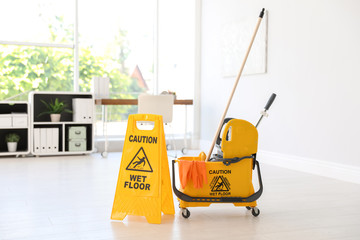 Phrase "CAUTION WET FLOOR" on safety sign and mop bucket with cleaning supplies, indoors © New Africa