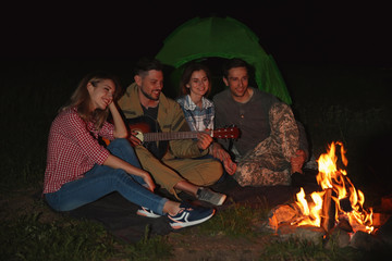 Young man playing guitar for friends near bonfire at night. Camping season