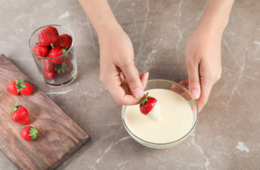Woman dipping ripe strawberry into bowl with white melted chocolate on table