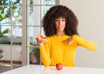 African american woman choosing between apple and pizza slice with surprise face pointing finger to...