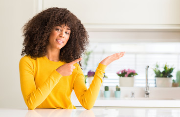 African american woman wearing yellow sweater at kitchen amazed and smiling to the camera while presenting with hand and pointing with finger.