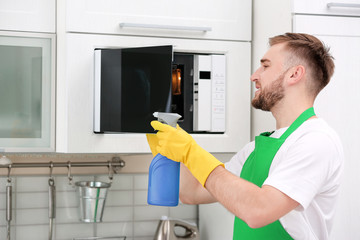 Young man in uniform cleaning microwave oven in kitchen