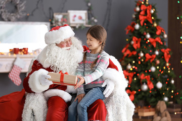 Fototapeta na wymiar Little girl with gift box sitting on authentic Santa Claus' lap indoors