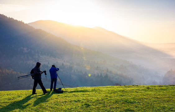 photographers on workshop at sunrise. capturing gorgeous scenery of mountainous area from the top of grassy hill. beautiful landscape with sun rising behind the forested mountain and foggy valley