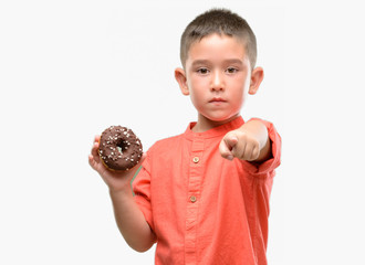 Dark haired little child eating doughnut pointing with finger to the camera and to you, hand sign,...