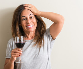 Middle age woman drinking red wine in a glass stressed with hand on head, shocked with shame and surprise face, angry and frustrated. Fear and upset for mistake.