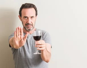 Selbstklebende Fototapeten Senior man drinking red wine in a glass with open hand doing stop sign with serious and confident expression, defense gesture © Krakenimages.com