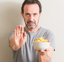 Senior man eating potato chips with open hand doing stop sign with serious and confident...