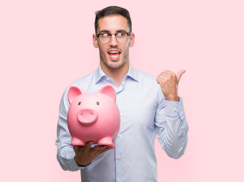 Handsome young man holding a piggy bank pointing with hand and finger up with happy face smiling