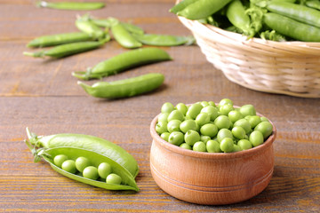 Fresh green peas in a wooden bowl and a basket of peas in the background on a brown wooden background