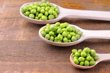 Fresh, green peas in different size wooden spoons on a brown wooden background