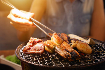 Barbecue meat and seafood on mesh grill of charcoal fire with women hand holding chopsticks. Korean or japanese traditional grilling style barbecue.