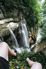 A guy in black shorts and sneakers sits on the edge of a rock in front of a large waterfall in the Agur gorge.